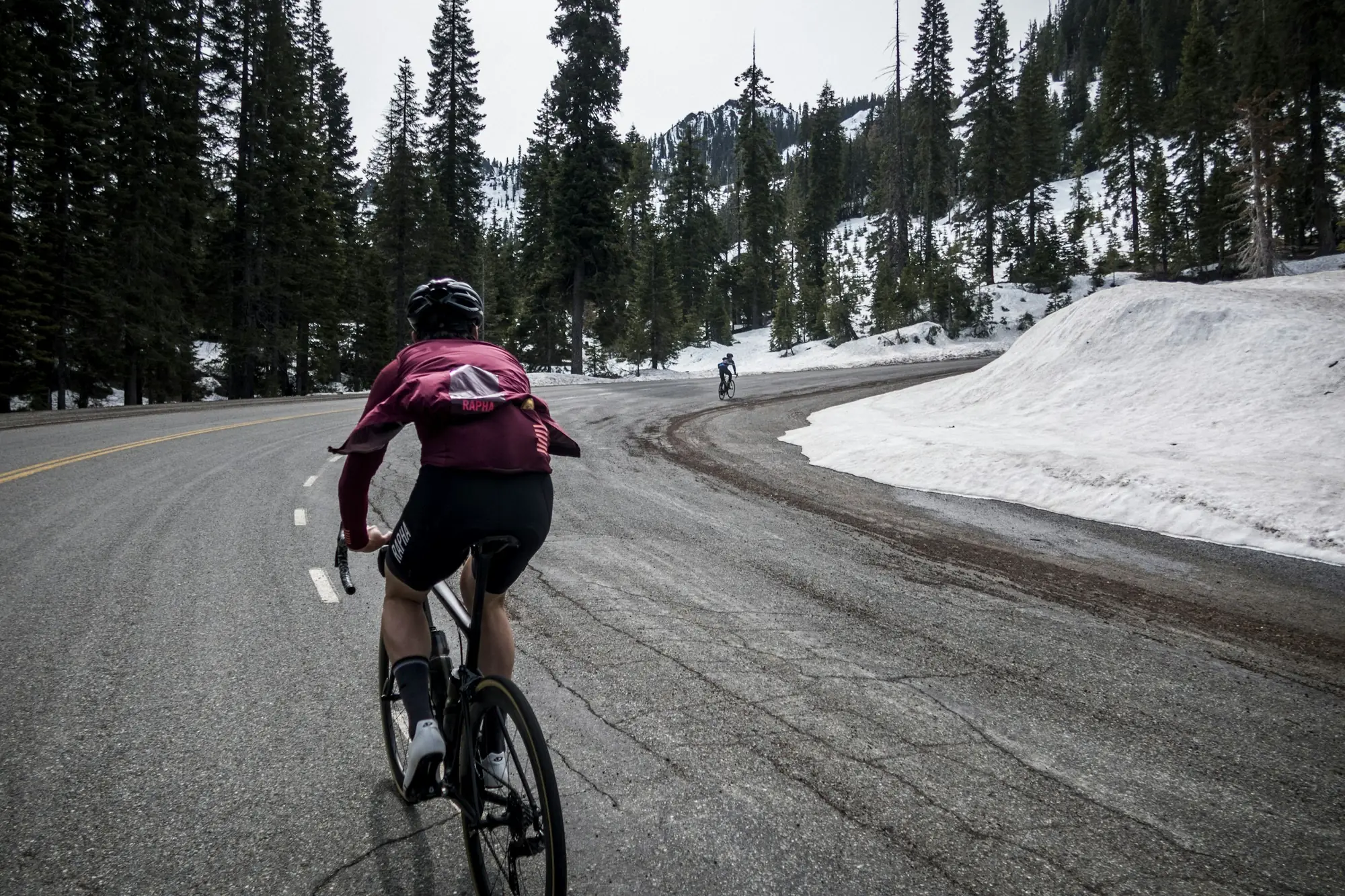 Un coureur de dos, maillot rouge, sur une route de col bordée de neige.